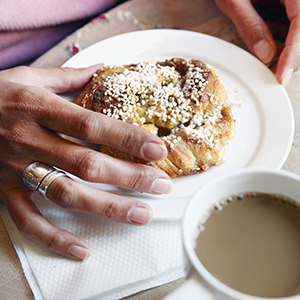 En hand greppar tag om en kanelbulle på ett fat. I förgrunden en kopp med kaffe.